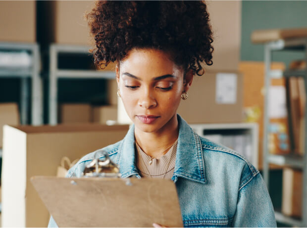 Mujer Revisando Un Inventario En Un Almacén, Sosteniendo Una Carpeta Con Hojas Y Rodeada De Cajas De Productos.